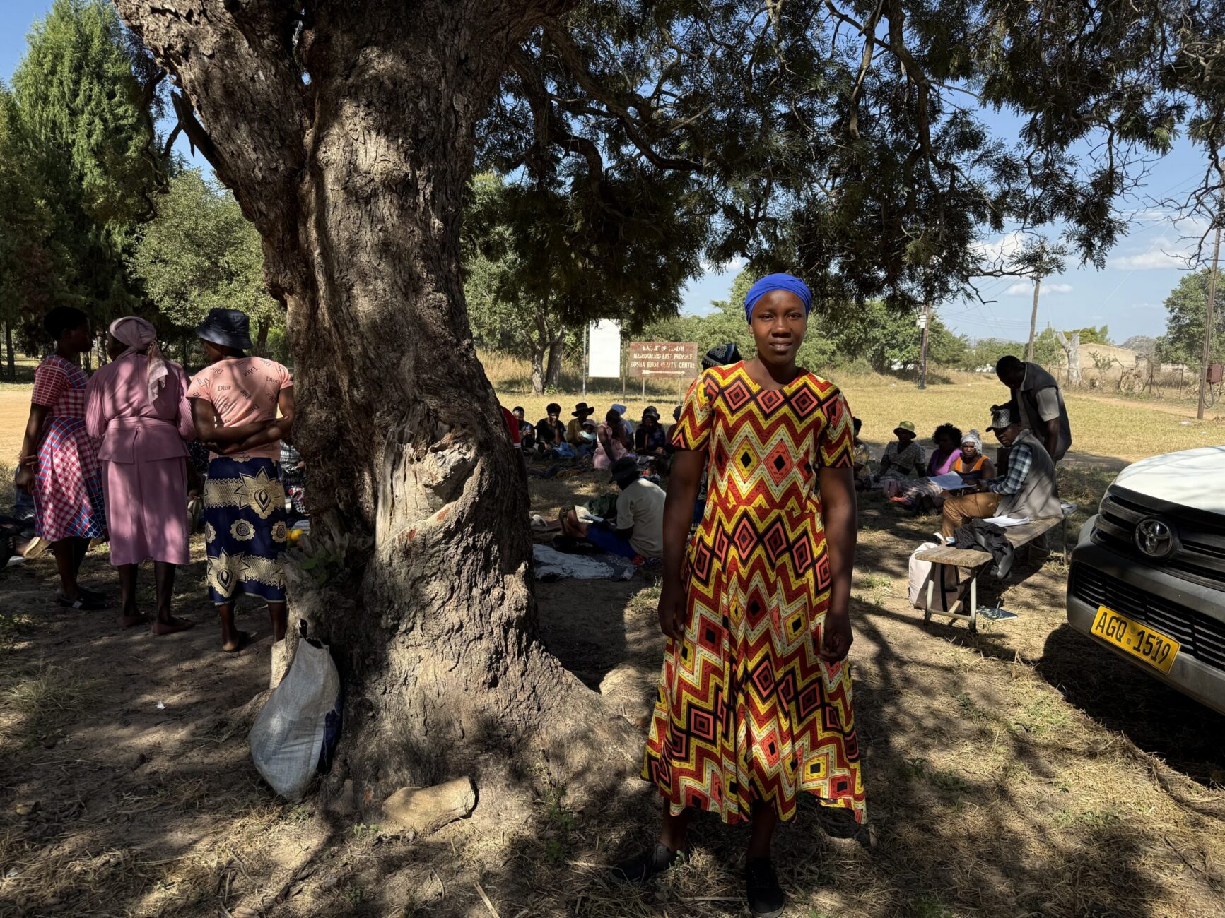 Wide shot of Lydia standing under a tree with women in the background